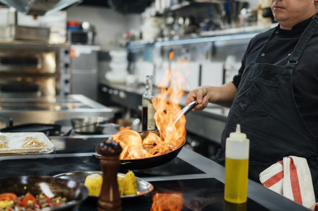 Chef preparing a fiery dish in a pot over open flames.